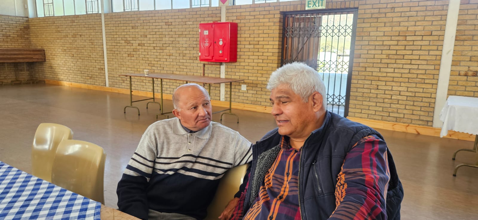 Two men sit close together in conversation inside the community hall, listening intently to one another.