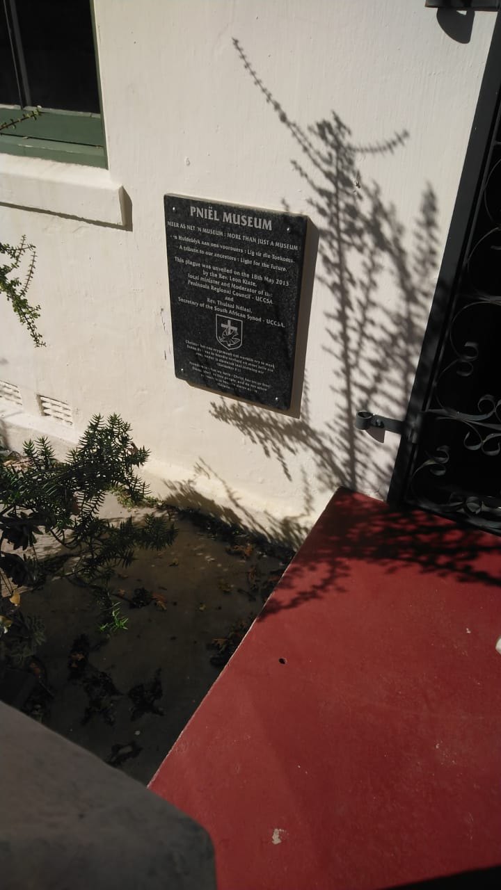 A black granite plaque mounted on a white wall reading Pniël Museum — Meer as net 'n museum / More than just a museum, dappled by leaf shadows.