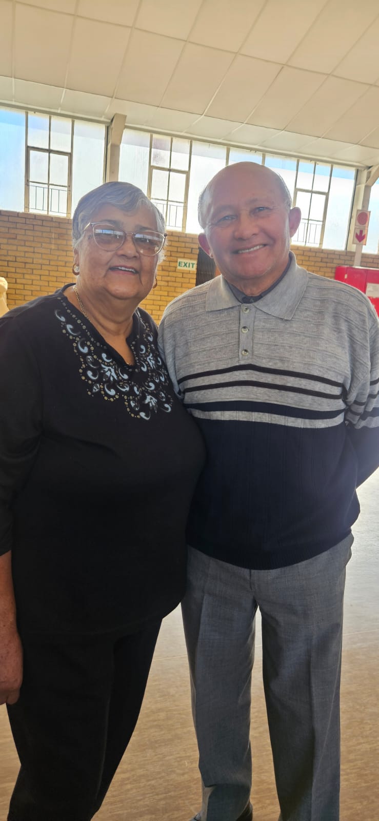 A smiling older woman and older man stand side by side in a hall, quietly proud.
