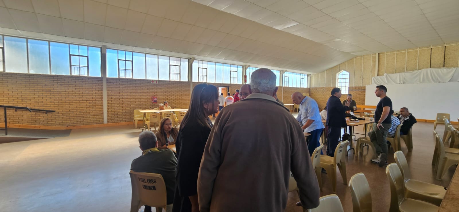 Community members gathering inside a bright church hall, standing and chatting among tables and chairs.