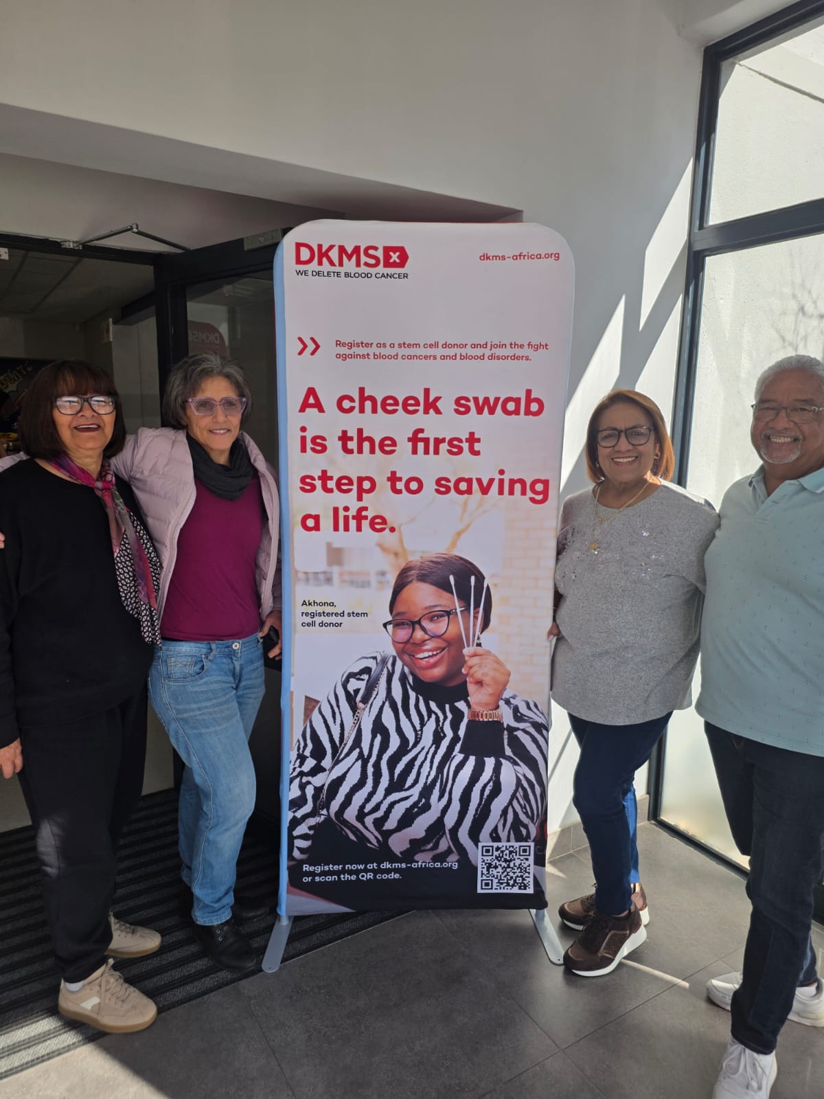 Four community members standing beside a DKMS Africa banner reading 'A cheek swab is the first step to saving a life.'