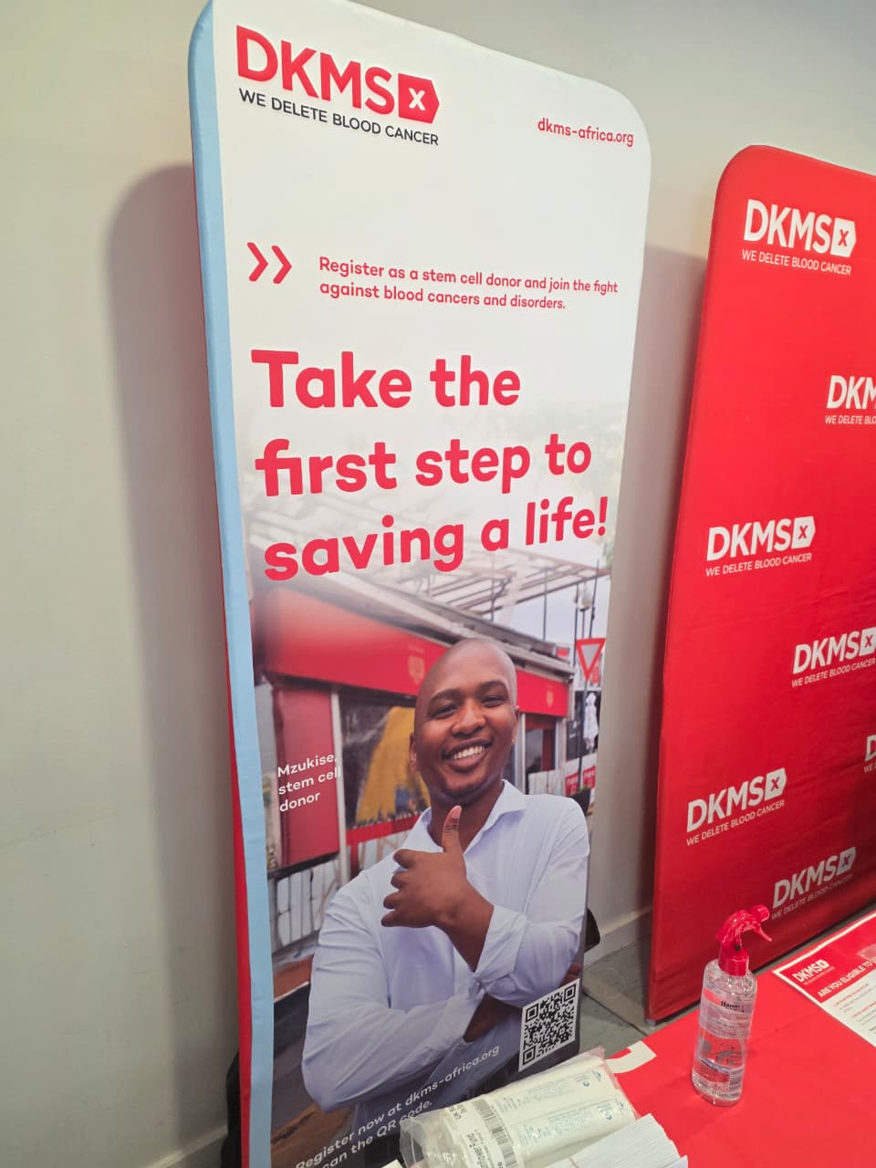 A DKMS Africa banner reading 'Take the first step to saving a life' beside a table of swab kits.