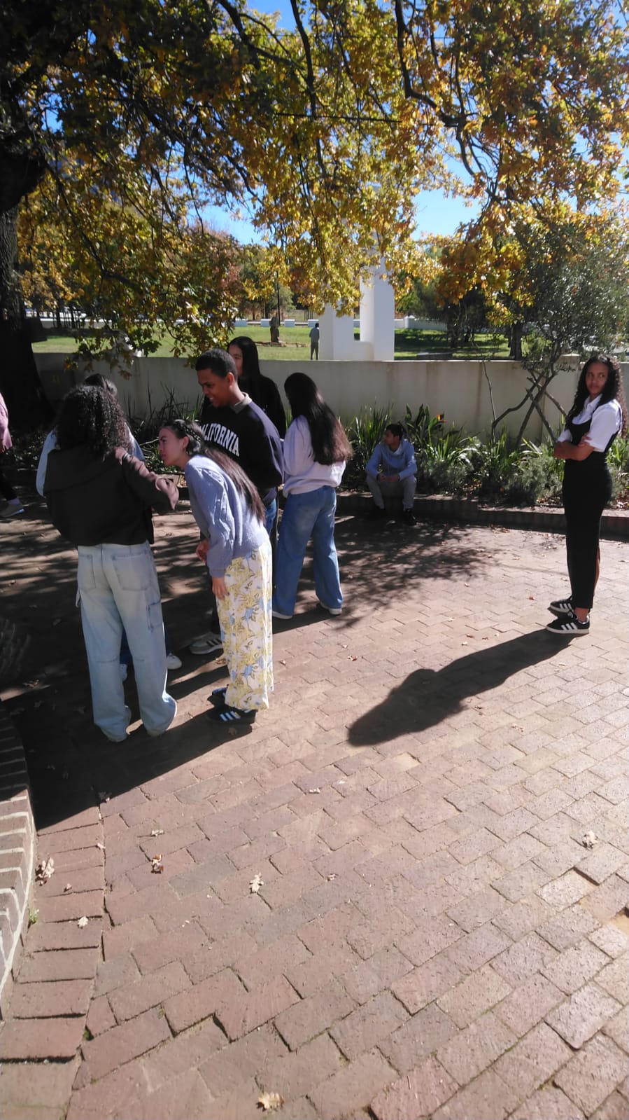 Teenagers in casual clothes stand talking on a brick courtyard beneath autumn-yellow oak branches, a white monument visible on the lawn beyond a low wall.