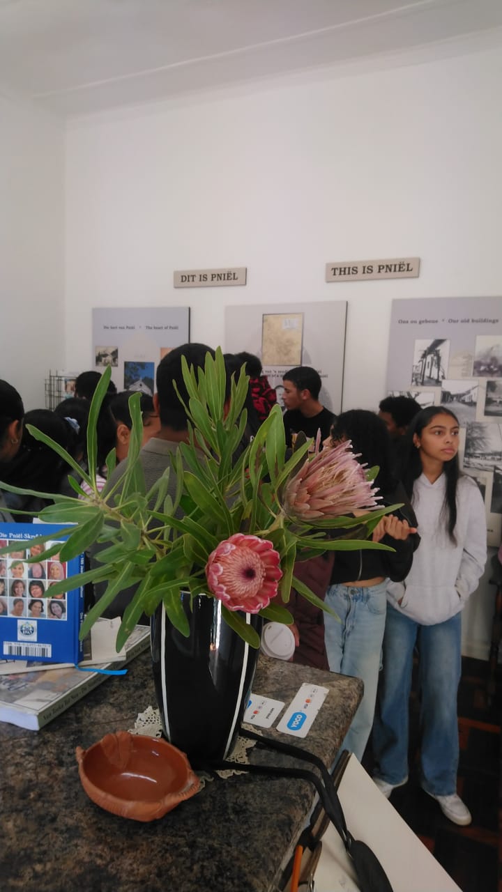 A tall vase of king proteas on a granite counter inside the museum, with visitors and exhibition panels titled Dit is Pniël and This is Pniël in the background.