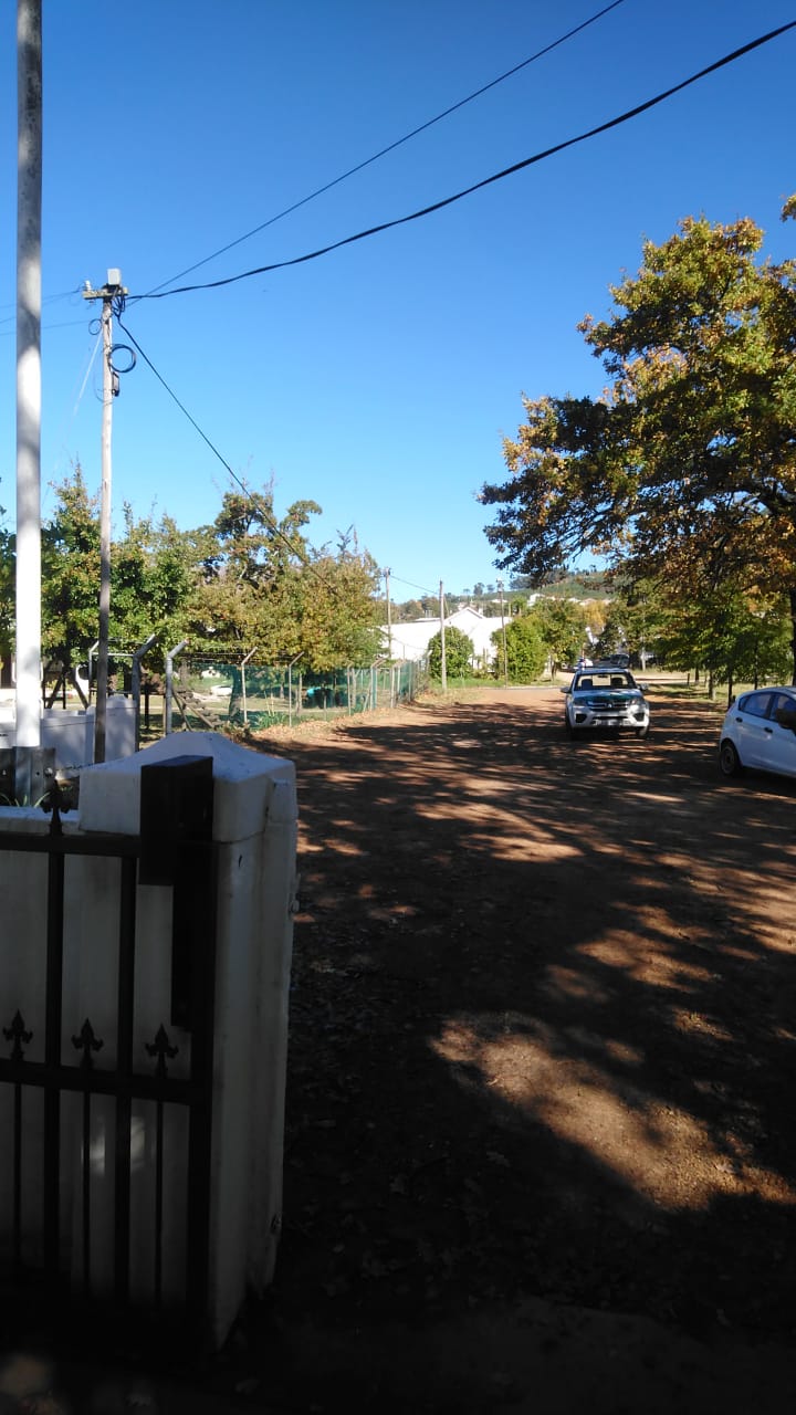 A white pillar and wrought-iron gate opening onto a dirt path shaded by oak trees, with cars parked further along.