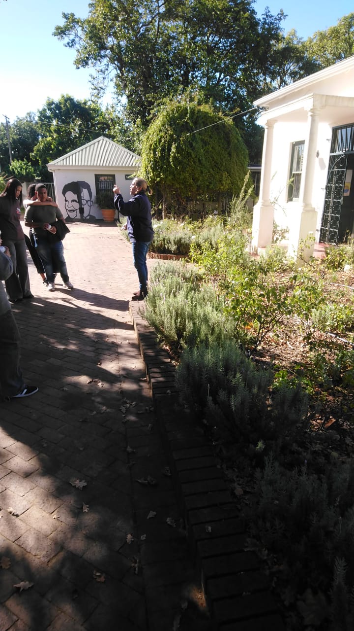 A woman in a black jacket points toward a painted black-and-white portrait mural on a cottage wall while listeners stand to one side of a lavender-edged path.